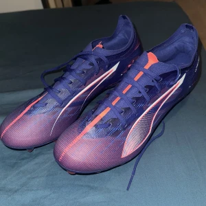 Puma Ultra fotbollsskor blå/röd - Säljer ett par Puma Ultra fotbollsskor i storlek 41 som är helt nya, oanvänd. Skorna är mörkblå med röda och vita detaljer, har snörning och en platt sula med dobbar för gräsplan. Ovandelen är tillverkad i syntetmaterial och har en smal, sportig form med rund tå. Perfekt för dig som vill ha speed på planen. Är normal i storlek