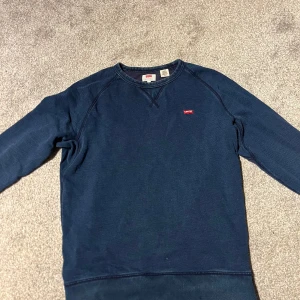Mörkblå sweatshirt från Levi's - Säljer en mörkblå sweatshirt från Levi's med klassisk crewneck och långärmad design. Tröjan har ett litet rött Levi's-märke på bröstet och är tillverkad i mjuk bomull. Perfekt för chill dagar och enkel att matcha med jeans eller joggers.