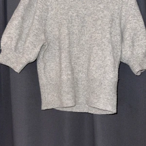 Grå stickad tröja med puffärm - Mysig grå stickad tröja med puffiga kortärmar och ribbade muddar. Tröjan har en rund halsringning och är i en ljusgrå ton som passar perfekt till jeans eller kjol. Materialet är mjukt och känns skönt mot huden, perfekt för kyliga dagar.💗