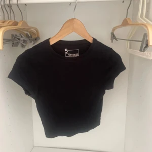 Svart croppad topp  - Svart croppad topp från FSBN Sister med rund hals och korta ärmar. Enkel och stilren design i mjuk bomull som passar perfekt till jeans eller kjol. Modellen är tight och ger en snygg siluett. Perfekt för dig som gillar minimalistisk stil.