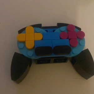 Ps 4 kontroll i Lego - Bilderna visar en färgglad spelkontroll byggd av LEGO-bitar, med tydliga knappar i gult och rosa samt svarta handtag. Perfekt för dig som gillar gaming och kreativt byggande! Ingen bok syns på bilderna.