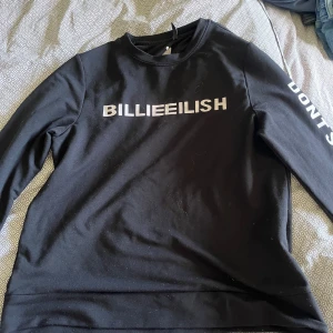 Svart Billie Eilish tröja från ONLY - Svart långärmad tröja från ONLY med Billie Eilish-tryck i vitt på bröstet och texten 'DON'T SMILE AT ME' längs ena ärmen. Tröjan har rund hals och är gjord i mjuk bomull. Ikonisk Billie Eilish-symbol tryckt på ärmen och insidan av halsen. Perfekt för fans av Billie Eilish.