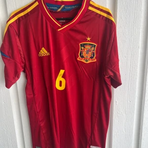 Spaniens Iniesta #6 Adidas matchtröja - Säljer en officiell röd Spanien fotbollströja från Adidas med gula detaljer och nummer 6 på bröstet och ryggen. Tröjan har korta ärmar, broderat landslagsmärke och Iniesta-tryck på ryggen. Materialet är lätt och andas, perfekt för match eller träning.