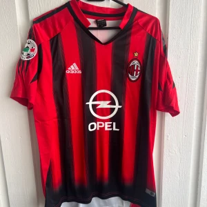 AC Milan Kaka' 22 Adidas fotbollströja - Säljer en klassisk AC Milan fotbollströja med Kaka' och nummer 22 på ryggen. Tröjan är röd och svart med vertikala ränder, Adidas-logga och Opel-sponsor på bröstet. V-ringad krage och korta ärmar, tillverkad i lätt polyester som andas. Perfekt för fans av Serie A och retrostil.