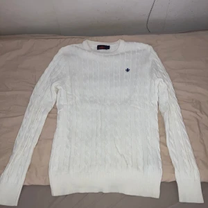 Vit kabelstickad tröja från Morris - Säljer en vit kabelstickad tröja från Morris med rund hals och diskret blå logga broderad på bröstet. Tröjan har långärmade ribbade muddar och är gjord i mjuk bomull, perfekt för dig som gillar klassisk och clean stil.