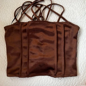 Brun korsett-topp från Divided XS - Snygg brun korsett-topp från Divided med glansig finish och smala axelband. Toppen har cool snörning i ryggen som ger en edgy vibe och markerade sömmar framtill för extra struktur. Perfekt att styla med jeans eller kjol för en trendig look.