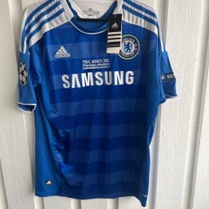 Chelsea FC Drogba 11 Finaltröja - Chelsea FC matchtröja från Champions League-finalen 2012 med Drogba 11 på ryggen. Adidas, blå med vita detaljer och ränder, officiella klubbmärken och Samsung-tryck på bröstet. Kortärmad, lätt och ventilerande material, perfekt för fotbollsfans.
