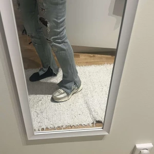 Grå slitna bootcut jeans med slits - Säljer ett par grå jeans med bootcut passform och råa slitsar vid bensluten. Byxorna har flera stora slitningar och hål längs benen för en edgy vibe. Jeansen är i klassisk denim och har en rak, något utsvängd siluett som ger en cool look.