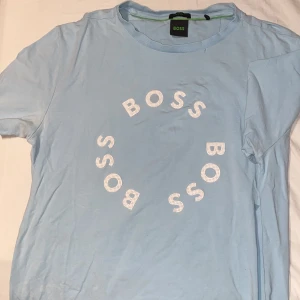 Ljusblå BOSS t-shirt med tryck - Fräsch ljusblå t-shirt från BOSS i mjuk bomull med stretch. Snyggt cirkulärt BOSS-tryck i vitt på bröstet. Klassisk rund hals och korta ärmar, perfekt för en chill och stilren look. Passar dig som gillar cleana och moderna plagg.