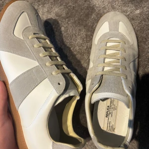 Maison Margiela Gats - Maison Margiela sneakers i stilren design med grå mockadetaljer och vita paneler i skinn. Klassisk rund tå, platt sula i gummi och snörning framtill. Insidan är fodrad i mjukt skinn för extra komfort. Perfekt för dig som gillar minimalistisk och clean stil.