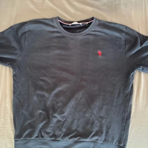 Mörkblå sweatshirt från U.S. Polo Assn. - Snygg mörkblå sweatshirt från U.S. Polo Assn. med klassisk rund hals och röd broderad logga på bröstet. Tröjan har ribbade muddar vid ärmslut och nederkant, vilket ger en schysst passform. Perfekt för dig som gillar stilren och enkel design.