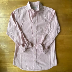 Rosa randig skjorta från The Shirt Factory - Slim fit skjorta från The Shirt Factory i storlek 38. Snyggt rosa och vitt randigt mönster, klassisk krage och långa ärmar. Skjortan har knappar framtill och är gjord i mjuk bomull som är enkel att tvätta och använda. Perfekt för dig som gillar stilrena detaljer.