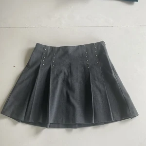 Grå plisserad kjol från Zara - Mörkgrå plisserad kortkjol från Zara med coola nitar framtill. Kjolen har en klassisk skoluniforms-vibe och är perfekt för dig som gillar en stilren look. Materialet känns mjukt och följsamt, och midjan är normal. Snygg att matcha med sneakers eller boots.