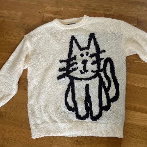 Vit stickad tröja med kattmotiv - Unik vit stickad tröja med ett stort svart kattmotiv både fram och bak. Tröjan har rund hals och långa ärmar, perfekt för dig som gillar coola och lekfulla prints. Materialet är mjukt och mysigt, passar dig som vill sticka ut med en rolig design.