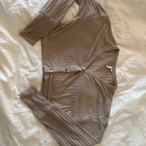 Beige ribbad croppad blus med knappar - Säljer en beige croppad blus med ribbad struktur och snygga knappar framtill. Modellen har lång ärm och v-ringad hals, perfekt att slänga över ett linne. Mjuk och skön i materialet, passar dig som gillar chill och stilren vibe.