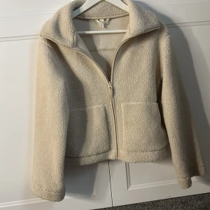 Beige teddyjacka med dragkedja XS - Mysig beige teddyjacka i storlek XS med två stora fickor framtill och dragkedja. Jackan har en bred krage och är helfärgad med långärmade ärmar. Perfekt för dig som gillar chill och comfy stil under kyliga dagar.