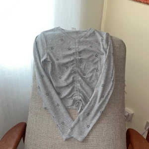 Grå långärmad topp med stjärnor Lindex❤️ - Grå långärmad topp från Lindex med svart stjärnmönster och snygg snörning framtill som ger en cool rynkad effekt. V-ringad och croppad passform, perfekt för dig som gillar detaljer. Mjuk och stretchig bomullskvalitet.🫶🏾🫶🏿🫶🏽🫶🏼