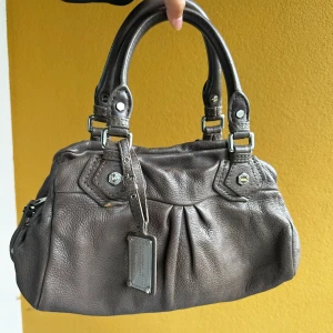 Vintage Brun handväska från Marc Jacobs - 100% koskinn, i gott skick. Det finns några små brister som visas på bilderna. Levereras med en avtagbar rem, så den kan användas både som axelväska och handväska. Storlek: 18 cm x 35 cm. Det är redan mitt lägsta pris, går tyvärr inte att pruta💕