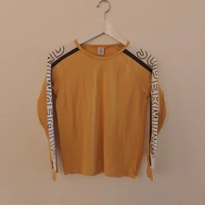 Lindex tröja  - Cool gul långärmad tröja från Lindex med svarta och vita detaljer på axlarna och texten 'SUPERMINDS' längs ärmarna. Skön passform och tillverkad i mjuk bomull, perfekt för dig som gillar streetstyle och vill sticka ut lite extra.