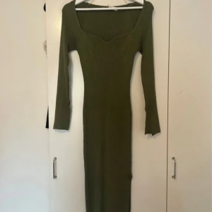 Olivgrön ribbad långklänning - Snygg olivgrön långklänning med ribbad struktur och figurnära passform. Klänningen har långärmade ärmar och en bred, fyrkantig urringning som ger en modern vibe. Perfekt för dig som gillar stilrena och trendiga plagg med enkel design.