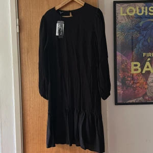 Svart långärmad klänning från Happy  Holly - Svart klänning från Happy ä Holly med rund halsringning och långa ärmar med resår vid ärmslut. Klänningen har en volang nedtill som ger en snygg och flowy siluett. Materialet är lätt och känns skönt mot huden. Perfekt för dig som gillar enkel men stilren design.