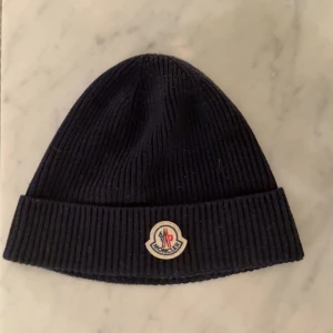Svart ribbad Moncler mössa - Snygg svart ribbstickad mössa från Moncler med klassisk uppvikt kant och ikonisk Moncler-logga framtill. Perfekt för dig som vill ha en stilren och exklusiv look. Mössan är mjuk och sitter skönt på huvudet, passar både till jacka och hoodie. Köpt ny i butik och det finns kvitto samt garanti som alla andra mina moncler plagg. Ny skicka