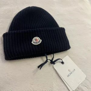 Svart ribbad mössa från Moncler - Snygg svart ribbstickad mössa från Moncler med uppvikt kant och klassisk logotyp framtill. Perfekt för kalla dagar och ger en clean look. Materialet är mjukt och stickat för extra komfort.
