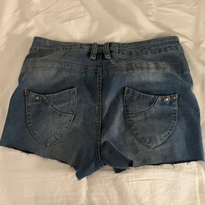 Blå jeansshorts med nitar - Snygga blå jeansshorts från Denim i storlek 36 med slim passform. Shortsen har råa kanter, fickor och coola nitar på fickorna fram och bak. Klassisk mid/ low waist och dragkedja framtill. 