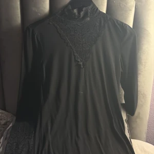 Svart spetsklänning från Soyaconcept - Snygg svart långärmad klänning från Soyaconcept med spetsdetaljer vid halsen och ärmsluten. Klänningen har en hög krage och ett elegant spetsmönster framtill som ger en cool och sofistikerad vibe. Perfekt för dig som gillar stilrena och detaljerade plagg.