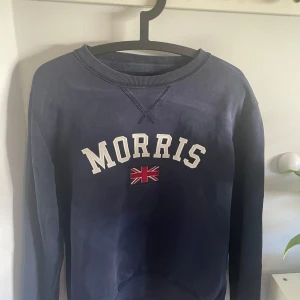 Morris tröja - Snygg mörkblå sweatshirt från Morris med broderad Union Jack-flagga och stora vita bokstäver på bröstet. Tröjan har rund hals och långa ärmar, tillverkad i mjuk bomull som är skön att bära. Perfekt för dig som gillar klassisk brittisk stil. Pyte litet hål som ni ser på  bild 3