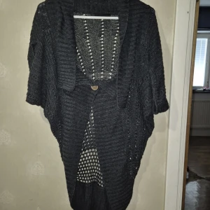 Mörkgrå stickad kofta med knapp - Säljer en mörkgrå stickad kofta med korta ärmar och snyggt hålmönster. Koftan har en stor träknapp framtill och en bred krage som ger en chill och avslappnad vibe. Perfekt att slänga över en t-shirt eller linne för extra lager. Passar dig som gillar mysiga och stilrena plagg.