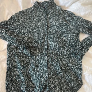 Grön mönstrad skjorta med knappar - Snygg grön skjorta med vitt prickigt mönster och knappar framtill. Skjortan har lång ärm och en avslappnad passform, perfekt till jeans eller kjol. Materialet känns lätt och luftigt, och kragen är klassisk. En cool detalj är det diskreta mönstret som gör skjortan unik.