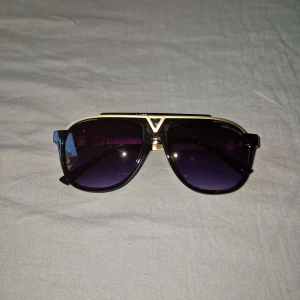 Louis Vuitton svart och guld solglasögon - Snygga solglasögon från Louis Vuitton med svart båge och guldiga detaljer. Glasen är mörka med en lila ton och har en cool V-formad detalj framtill. Perfekt accessoar för dig som vill sticka ut med exklusiv stil.