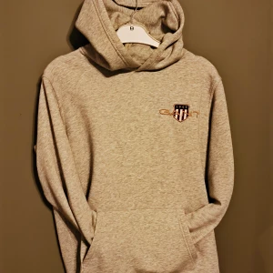 Grå hoodie från GANT med logga - Säljer en grå hoodie från GANT med broderad logga och emblem på bröstet. Tröjan har huva, magficka och långa ärmar. Mjuk och skön i bomullsmix, perfekt för chill dagar. Passar dig som gillar klassisk streetstyle och enkel design. Hoodien passar folk med S/M
