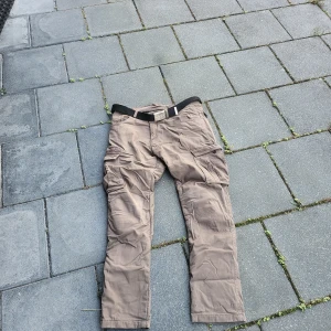 Beige cargopants(skydds byxor)från D-COURSE - Säljer ett par beige cargopants från D-COURSE med flera praktiska fickor på benen och medföljande svart bälte. Byxorna har en rak passform och är tillverkade av bomull och polyester, vilket gör dem både slitstarka och bekväma. Perfekta för dig som gillar funktionell stil.