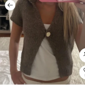 Brun stickad kofta - Säljer en brun stickad kofta med korta ärmar och en stor knapp framtill. Koftan har en v-ringad öppning och ribbade muddar vid ärmslut och nederkant. Perfekt att slänga över en t-shirt för en chill look.