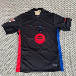 FC Barcelona Lamine Yamal Nike tröja - Snygg FC Barcelona matchtröja med Lamine Yamal och nummer 19 på ryggen. Svart bas med röda och blå detaljer på sidorna, röd Spotify-logga och klubbmärke framtill. Tillverkad i lätt och ventilerande polyester, kortärmad och med modern krage. Perfekt för dig som älskar Barca.