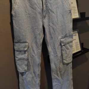 Ljusblå cargopants i jeansmaterial - Säljer ett par ljusblå cargopants i jeansmaterial med breda ben och stora fickor på sidorna och bak. Perfekta för dig som gillar streetstyle och vill ha mycket plats för prylar. 
