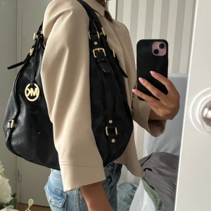 Svart axelväska Michael Kors skinn - Snygg svart axelväska från Michael Kors i mjukt skinn med guldfärgade detaljer och MK-logga. Väskan har flera fack, dragkedja och justerbara remmar. Insidan är fodrad med MK-mönster. Perfekt storlek för allt du behöver ha med dig.