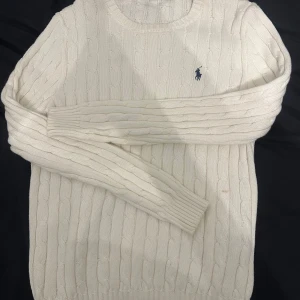 Vit stickad tröja från Ralph Lauren - Säljer en klassisk vit stickad tröja från Ralph Lauren med kabelstickat mönster och rund hals. Tröjan har långärmade ribbade muddar och den ikoniska blå Polo-loggan broderad på bröstet. Perfekt för dig som gillar stilren och tidlös design.