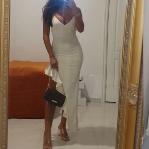 Champagnefärgad långklänning med volang - Säljer en elegant champagnefärgad långklänning från Zara med smala axelband och v-ringad urringning. Klänningen har en asymmetrisk volangdetalj längs sidan och är figurnära med en snygg slits. Perfekt för dig som vill sticka ut med stil.