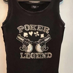 Poker Legend svart paljettlinne - Svart ärmlöst linne från Mystic med glittrande paljetter och stenar på framsidan. Tryck med texten 'Poker Legend', Rå kant vid ärmhål och halsringning ger en edgy vibe. Perfekt för dig som gillar statement-plagg och bling.pris kan diskuteras!