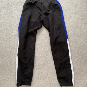 Svarta träningsbyxor från Lacoste - Snygga svarta träningsbyxor från Lacoste Sport med blå och vita paneler längs sidorna. Byxorna har elastisk midja, klassisk Lacoste-logga och text på benet. Materialet är lätt och syntetiskt, perfekt för sport eller chill. Passformen är normal och benen är raka med resår vid anklarna.