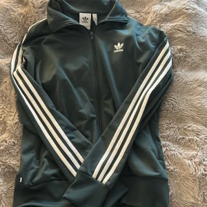 Adidas grön vindjacka med ränder - Snygg grön vindjacka från Adidas med klassiska vita ränder längs ärmarna och dragkedja framtill. Jackan har ståkrage och är tillverkad av polyester, perfekt för en sportig och chill stil. Ikonisk Adidas-logga på bröstet.
