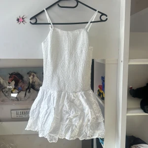 Vit kort klänning med spetsvolang - Supersöt vit klänning med smockad överdel och tunna axelband. Kjolen har två lager med volanger och broderad spetsdetalj som ger en romantisk vibe. Perfekt för sommarens alla tillfällen! Materialet känns lätt och luftigt.