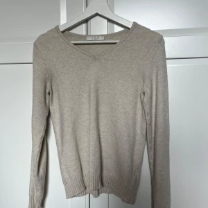 Beige stickad tröja från Vila - Snygg beige stickad tröja från Vila i storlek XS. Tröjan har v-ringad hals, långa ärmar och ribbade muddar vid ärmslut och nederkant. Perfekt för lager-på-lager och chill stil. Enkel att matcha med jeans eller kjol.