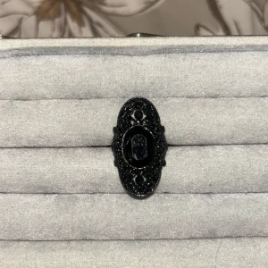 Svart oval statementring med sten - Snygg svart ring med stor oval form och detaljerad ram. Ringen har en glansig svart sten i mitten och filigranliknande mönster runt om som ger en cool vintagekänsla. Perfekt för dig som gillar att sticka ut med dina accessoarer.