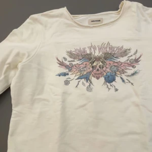 Vit långärmad tröja Zadig & Voltaire - Säljer en vit långärmad tröja från Zadig & Voltaire som orginelt kostar 1250 med coolt tryck framtill i rosa, blått och beige. Trycket har blommor, döskalle och vingar som ger en edgy vibe. Tröjan har rund hals och är gjord i mjuk bomull. Perfekt för dig som gillar statement-plagg! Som är liten i storleken är lite liten för mig och jag är 13 år🩷
