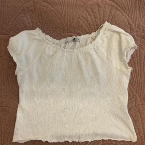 Vit blus med rynkad struktur från lager 157 - Supersöt vit blus från 57 i storlek M med rynkad, krispig struktur och volangkant vid halsringningen. Blusen har korta ärmar och en lätt croppad passform som ger en chill och fräsch vibe. Perfekt för att styla med jeans eller kjol för en trendig look.