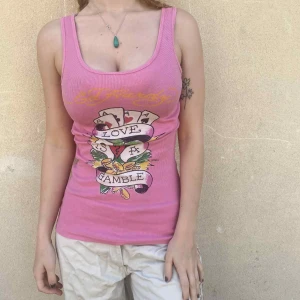 Rosa Ed Hardy linnetopp med tryck - Säljer en rosa ribbad linnetopp från Ed Hardy med ikoniskt "love is a Gamble" tryck framtill. Gult Ed Hardy-tryck på ryggen och nedtill. Slim fit och ärmlös, perfekt för dig som gillar statement-plagg. Helt äkta, använd ett fåtal gånger för ett par år sedan. 